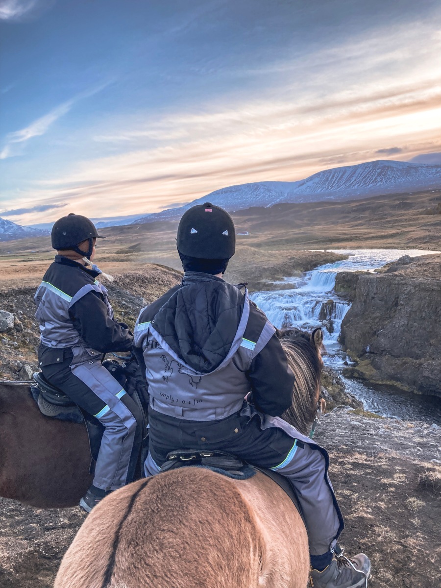 A young couple horse riding alongside waterfalls in Iceland, in a valley with mountainous scenery.
